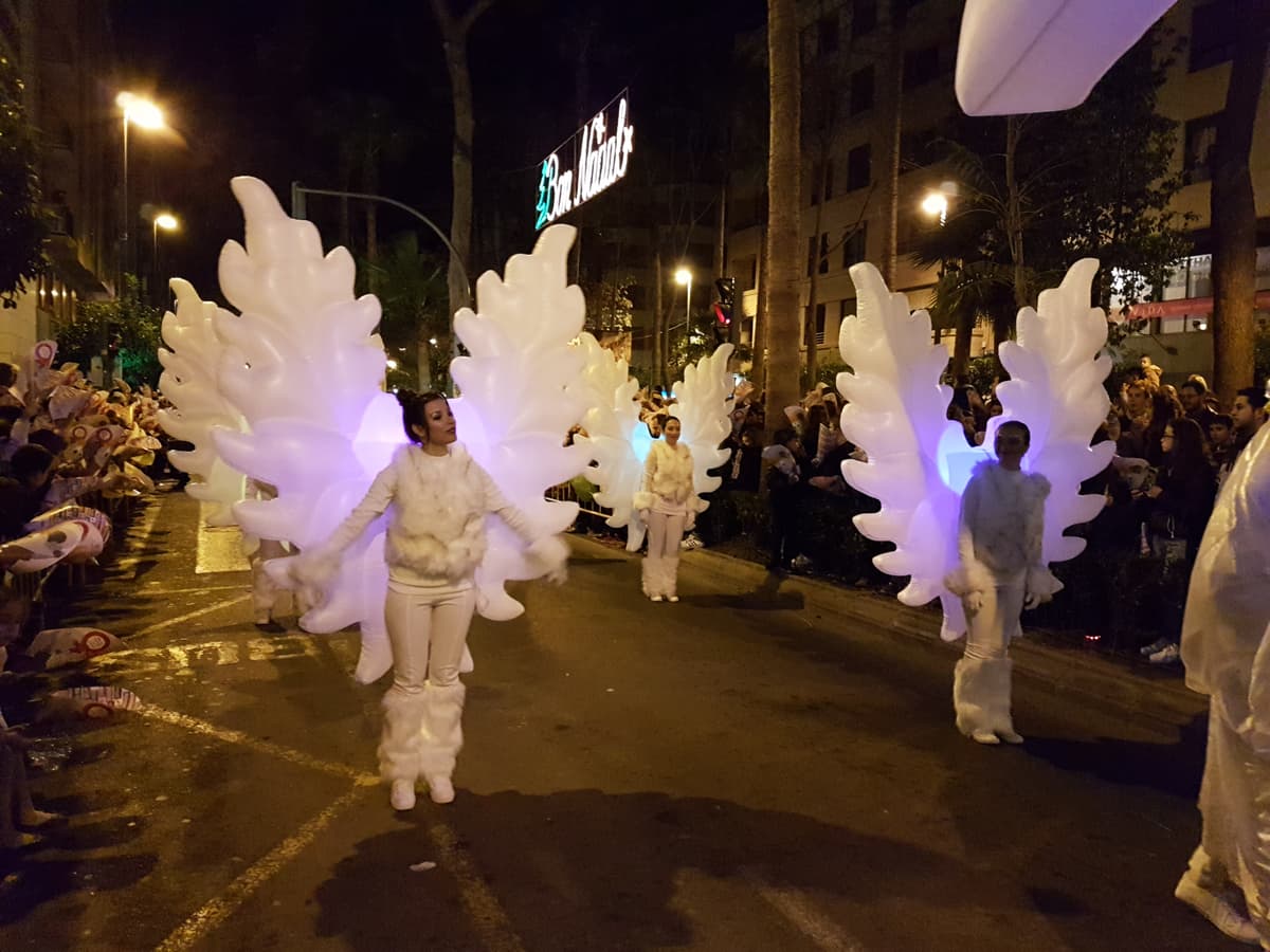 Bloque de ángeles con alas iluminadas avanzando por una calle en una cabalgata nocturna, con vallas y público