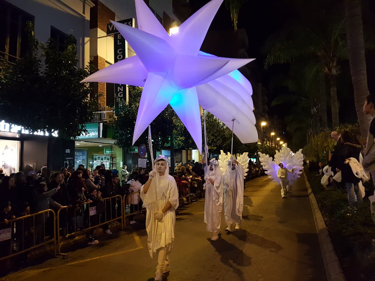 Gran estrella iluminada sobre un poste liderando un cortejo de figuras blancas y doradas en calle nocturna