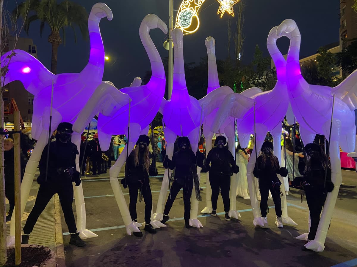 Fila de flamencos hinchables iluminados portados sobre pértigas por manejadores vestidos de negro