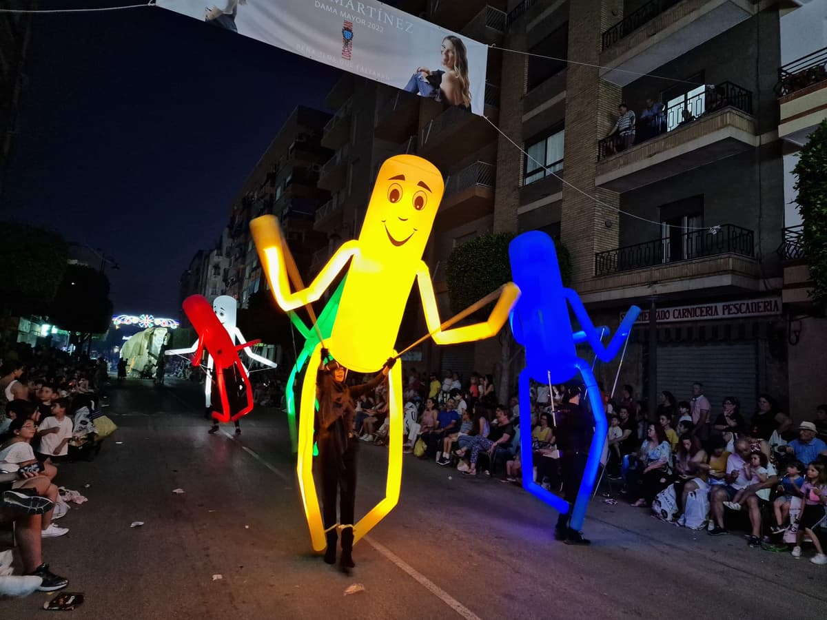 Marionetas gigantes de tubo en amarillo, azul, rojo y blanco avanzando por una calle nocturna con público sentado