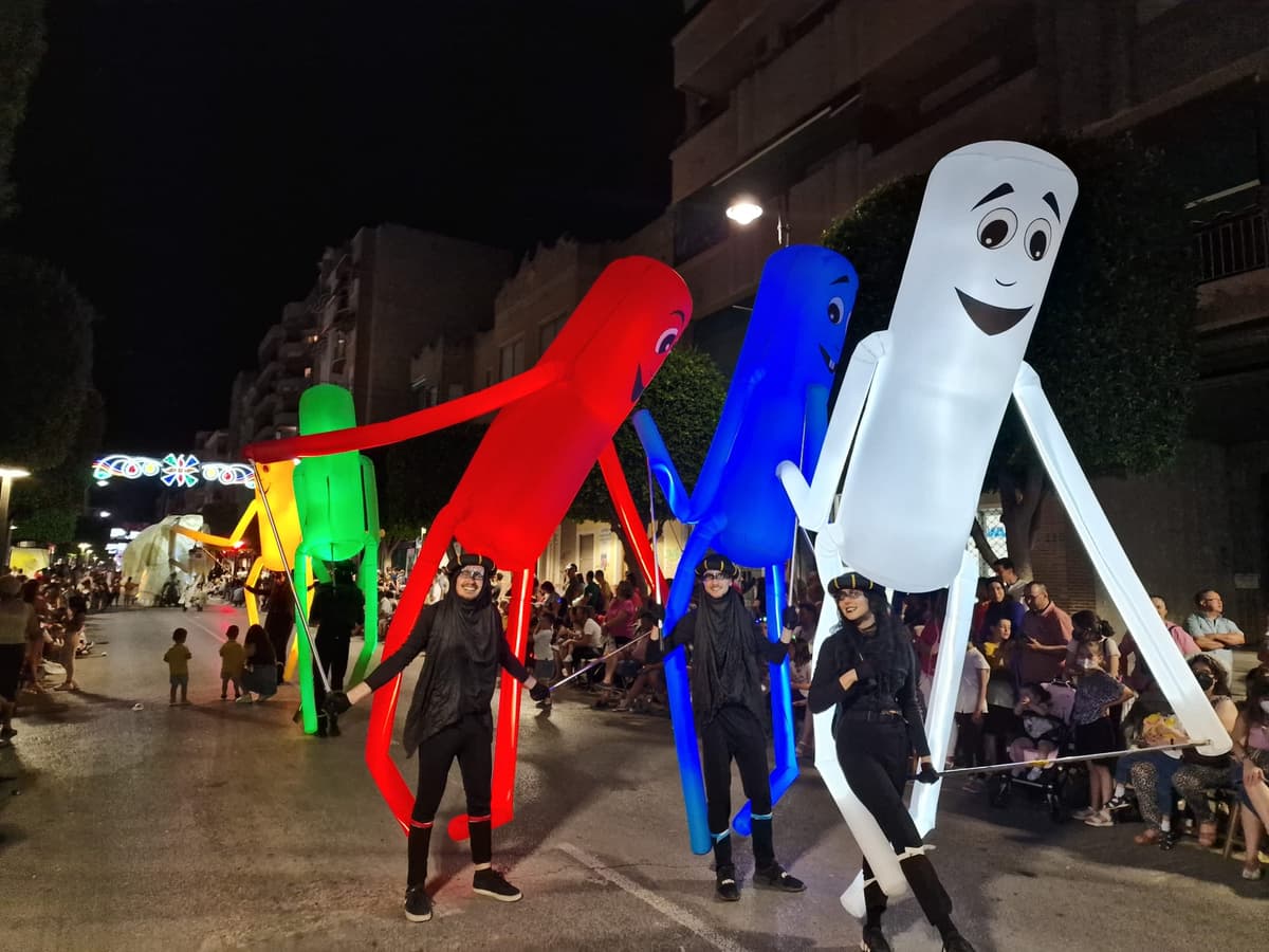 Cuatro marionetas gigantes de tubo en colores llevadas por sus operarios en una calle nocturna